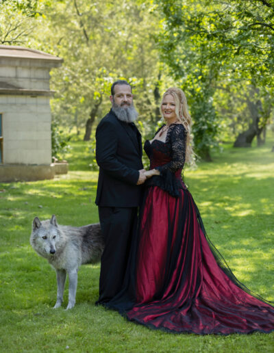 Couple in black and burgundy attire posing with a white wolf in a green park.
