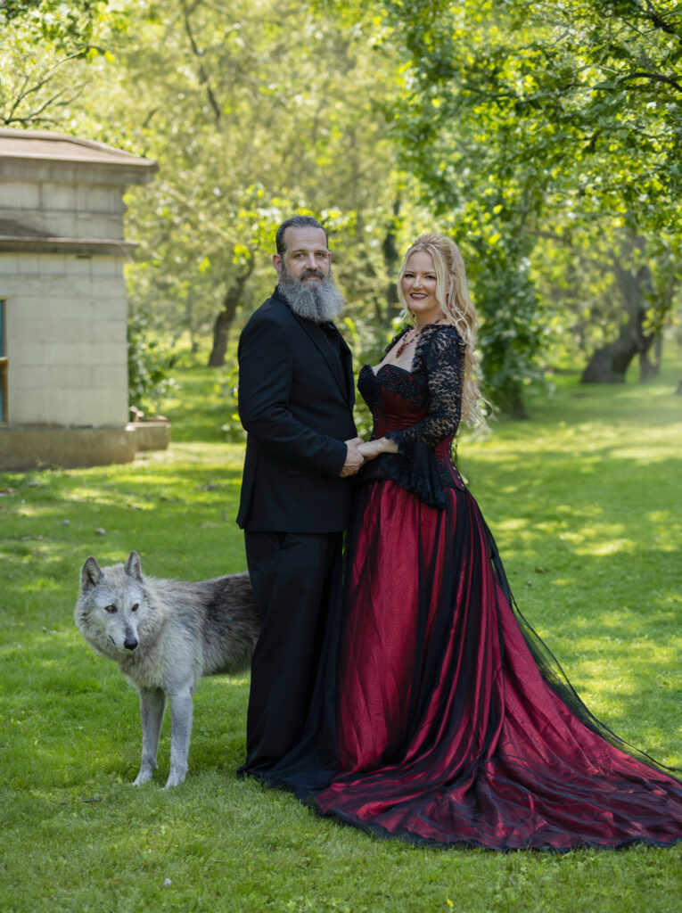 Couple in black and burgundy attire posing with a white wolf in a green park.