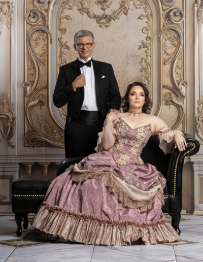 Man in a black tuxedo standing behind a woman seated in a pink ball gown.