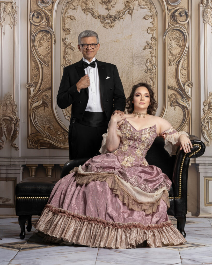 Man in a black tuxedo standing behind a woman seated in a pink ball gown.