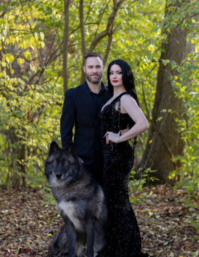 Couple in dramatic black clothing posing together in a sunlit wooded setting.