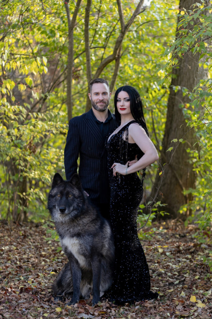 Couple in dramatic black clothing posing together in a sunlit wooded setting.