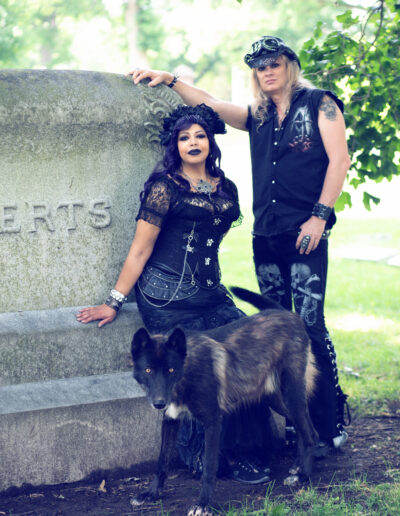 Gothic couple in dark blue and black attire posing together in a cemetery.