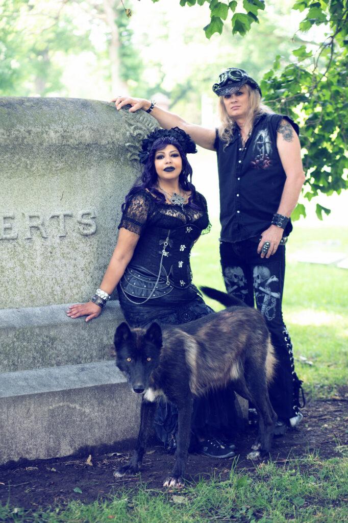 Gothic couple in dark blue and black attire posing together in a cemetery.