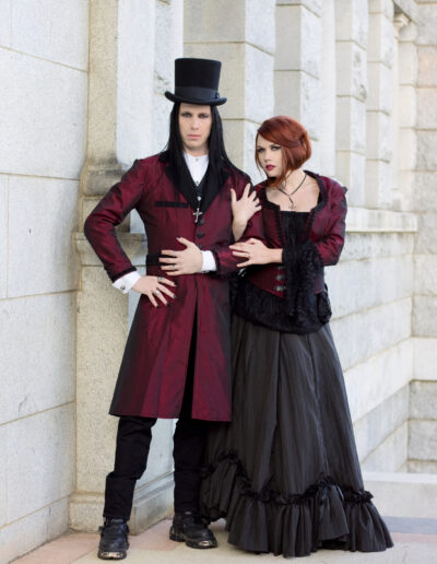 Couple in red and black Victorian-inspired clothing posing beside a stone building.