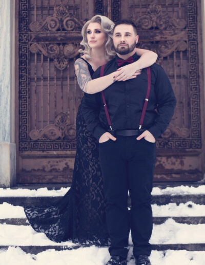Gothic couple in black and burgundy attire posing together before ornate doors.