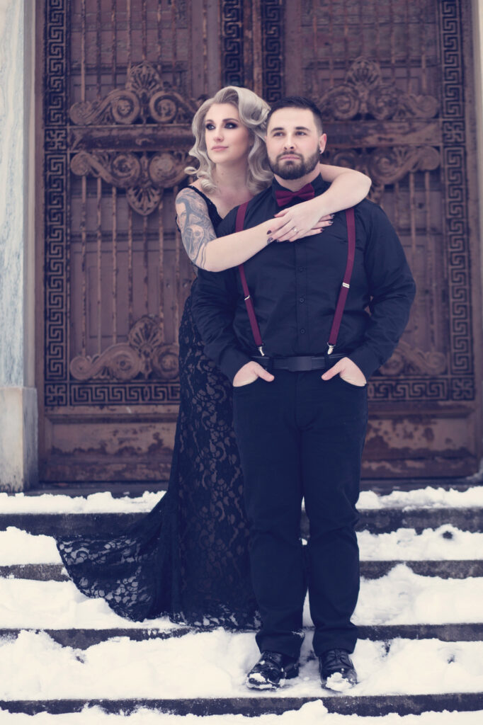 Gothic couple in black and burgundy attire posing together before ornate doors.