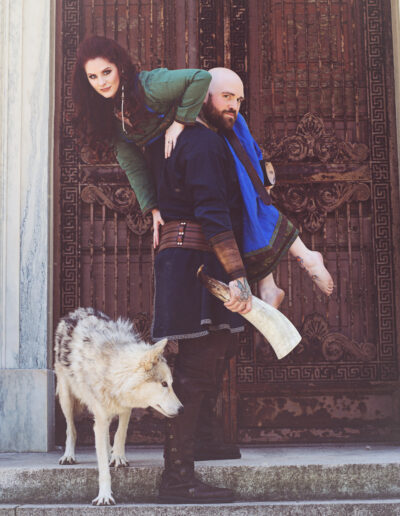 Fantasy couple in jewel-toned costumes posing together against a textured brick wall.