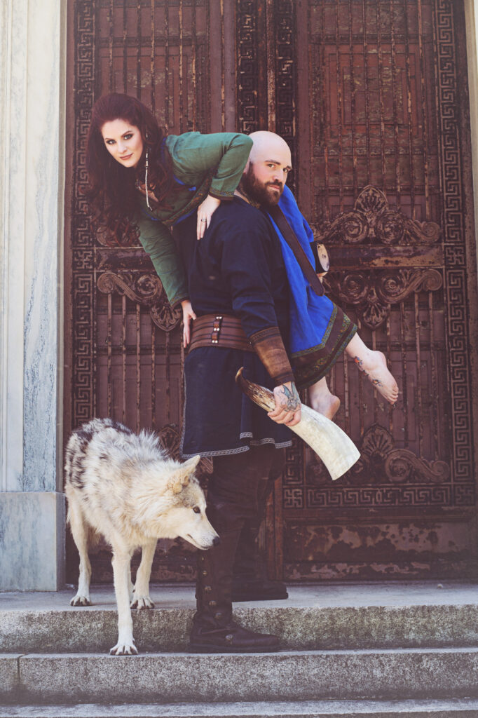 Fantasy couple in jewel-toned costumes posing together against a textured brick wall.