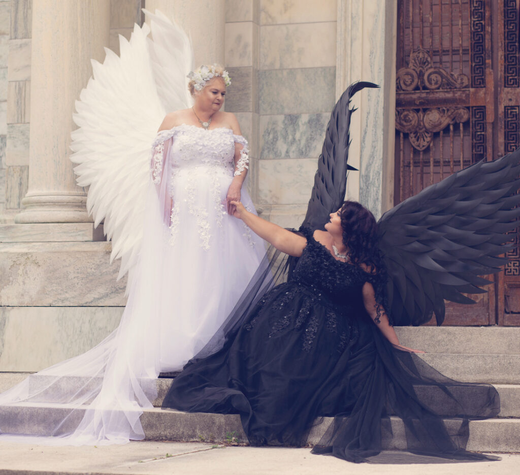 Two women portraying light and dark angels with contrasting feathered wings.