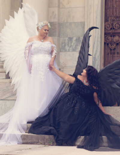 Two women portraying light and dark angels with contrasting feathered wings.