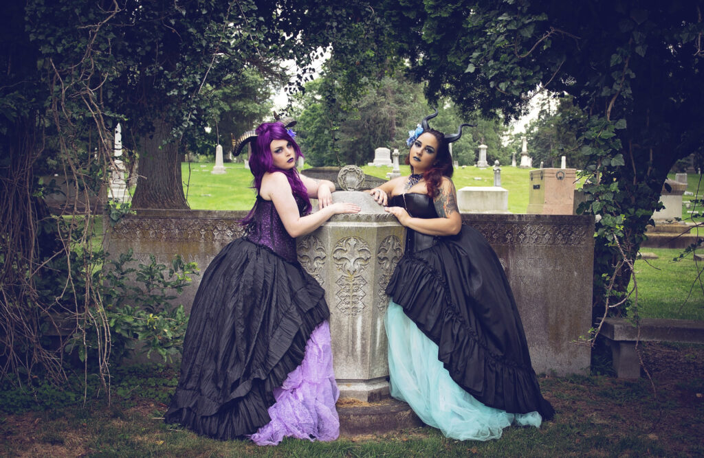 Two women in flowing dark gowns posing beside a stone monument in a wooded cemetery.