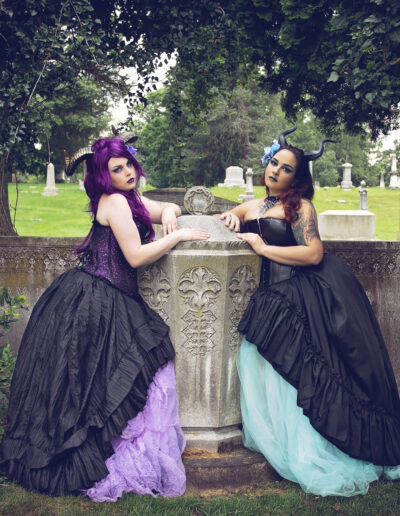 Two women in flowing dark gowns posing beside a stone monument in a wooded cemetery.