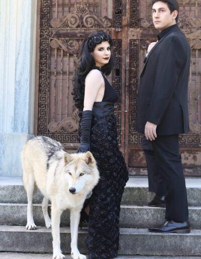 Gothic couple in black posing with white wolves outside an ornate stone building.