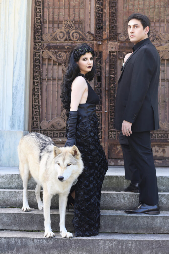 Gothic couple in black posing with white wolves outside an ornate stone building.