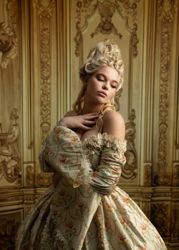 Blonde woman in a cream floral period gown posed against ornate gold paneling.