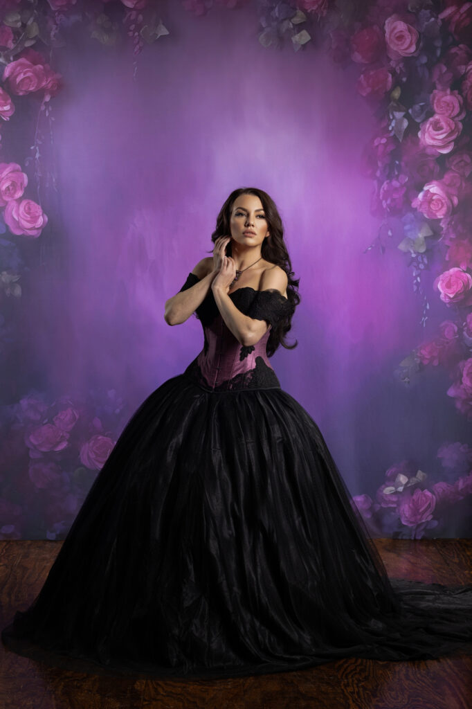 Woman in a black and purple corseted Victorian gown surrounded by purple roses.