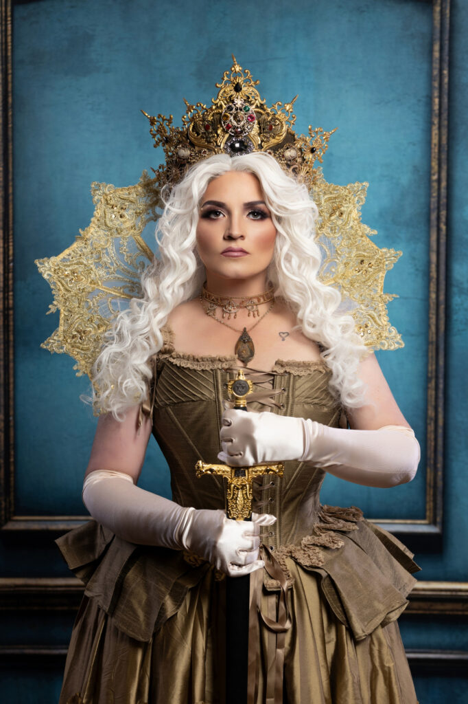 White-haired woman wearing a jeweled crown, corseted period gown and white gloves.