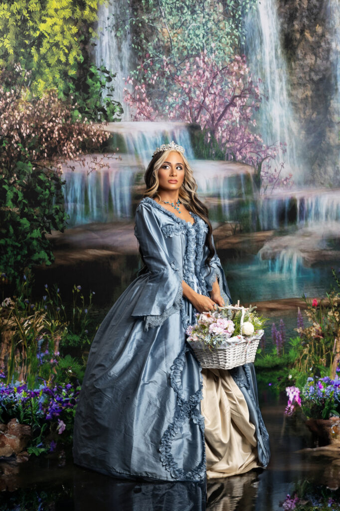Woman in a blue Victorian gown carrying a flower basket through a waterfall garden.