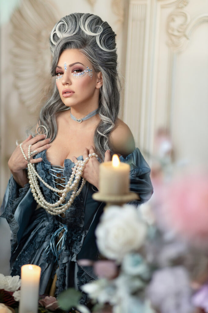 Silver-haired woman in a blue corseted Victorian gown holding pearls beside candlelight.