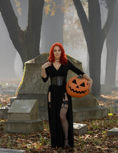 Red-haired gothic woman seated on a tomb with a jack-o’-lantern