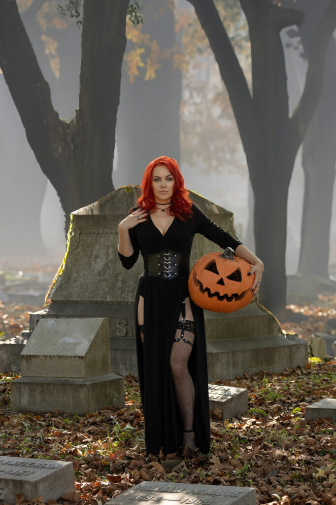 Red-haired gothic woman seated on a tomb with a jack-o’-lantern