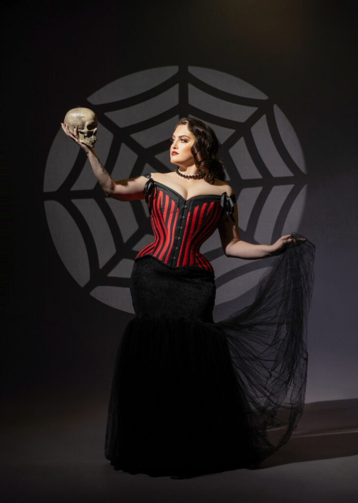 Gothic woman in a red-and-black corset posed against a spiderweb backdrop