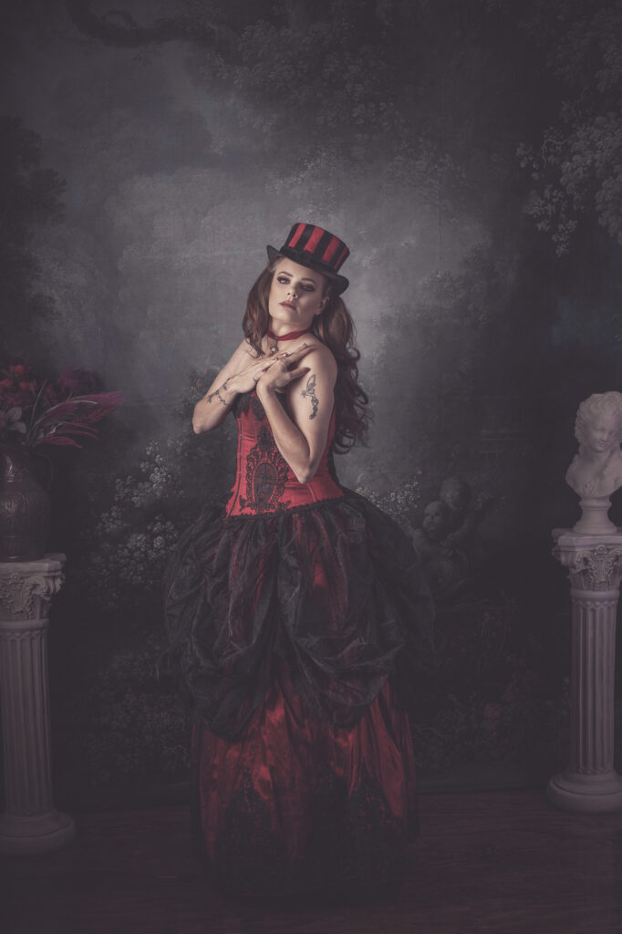 Woman in a red and black steampunk gown with a striped top hat in a moody studio.