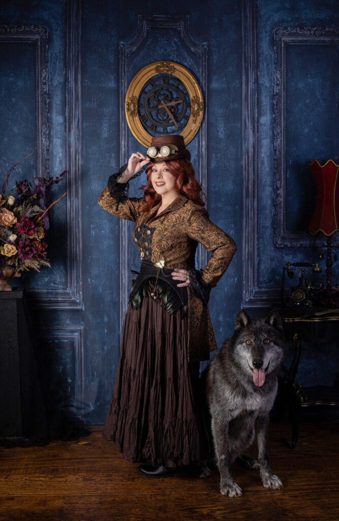 Woman in brown steampunk attire and goggles posing with a dog in an ornate blue parlor.