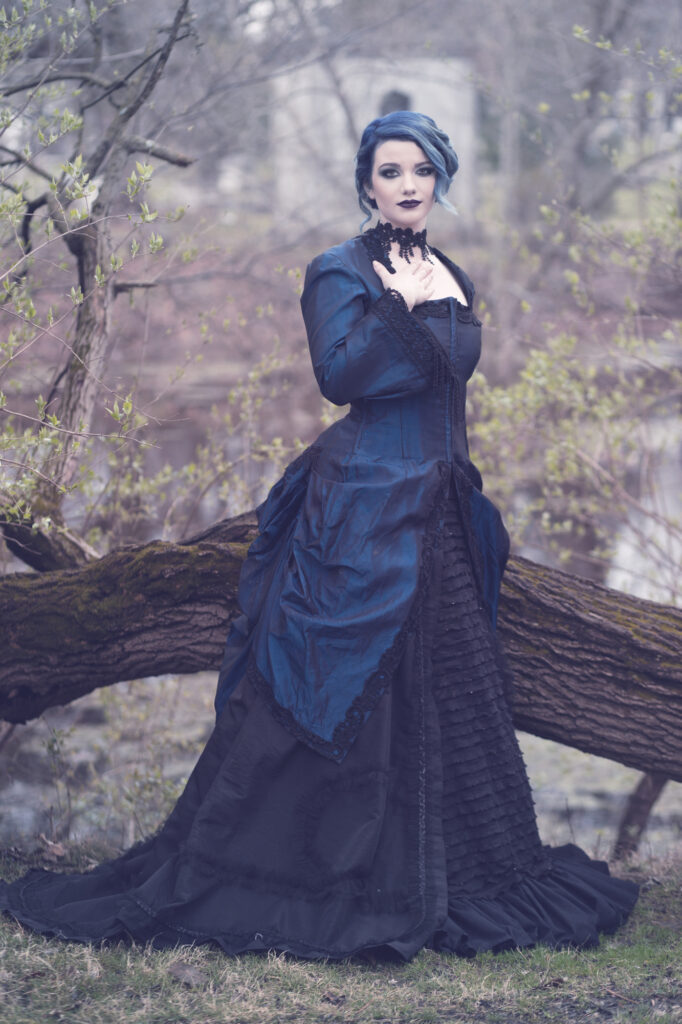 Woman in a dark blue Victorian gown posing on a fallen tree in a misty woodland.
