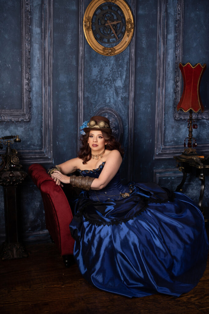 Woman in a deep blue Victorian ball gown seated on a red chaise in an ornate room.