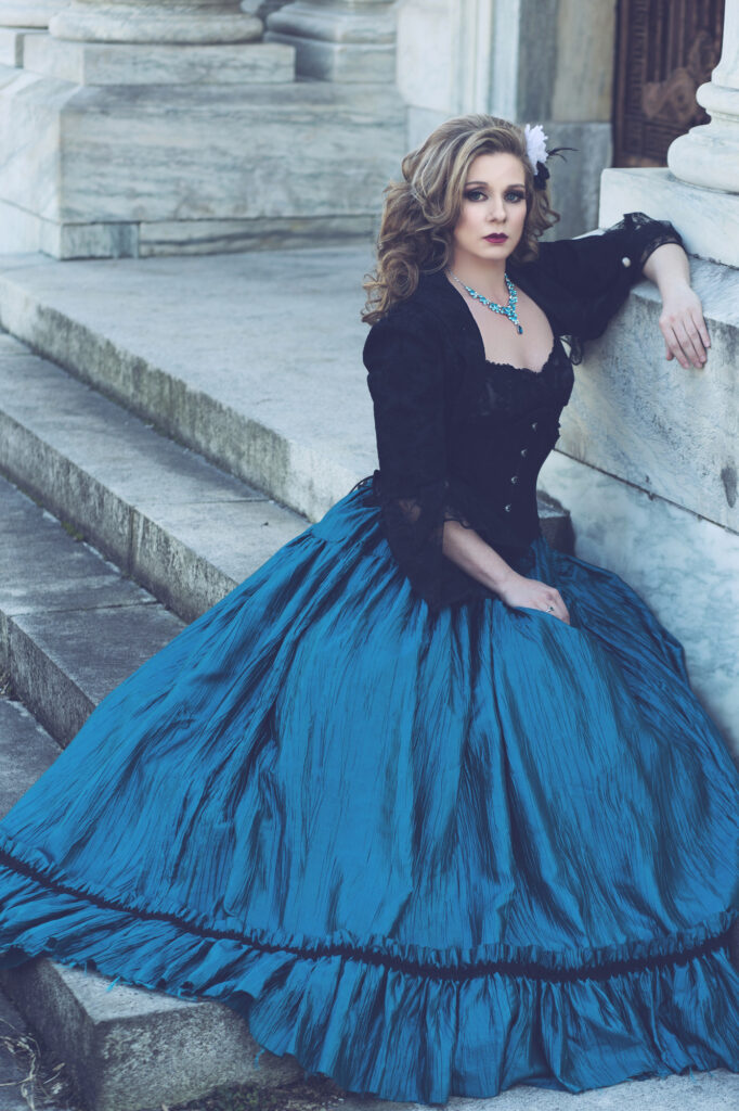 Woman in a black Victorian bodice and dramatic blue skirt seated on stone steps.