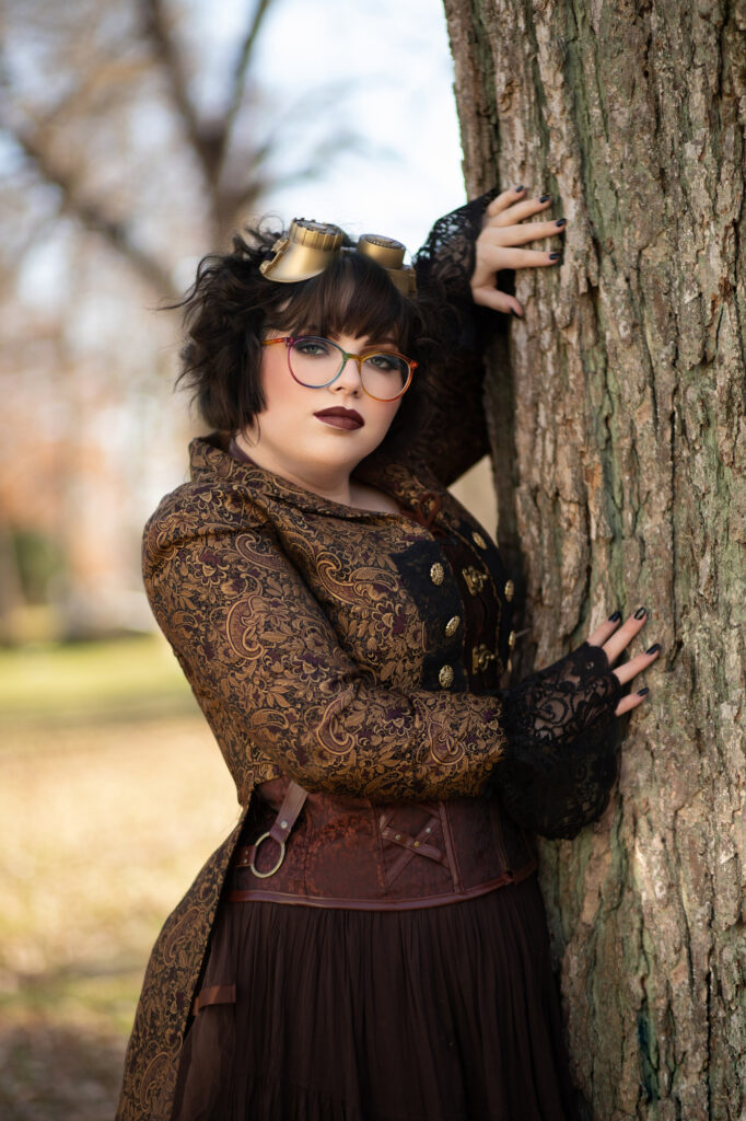 Woman wearing goggles, glasses and a brown steampunk corset while leaning against a tree.