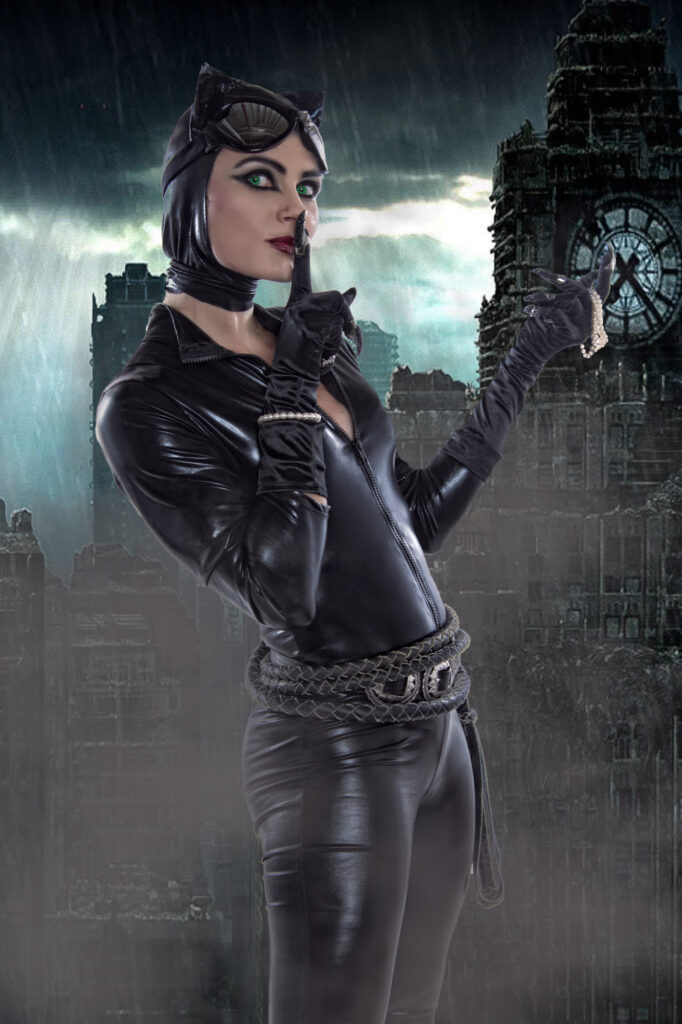 Woman in a black leather hooded costume posing in a misty wooded setting.