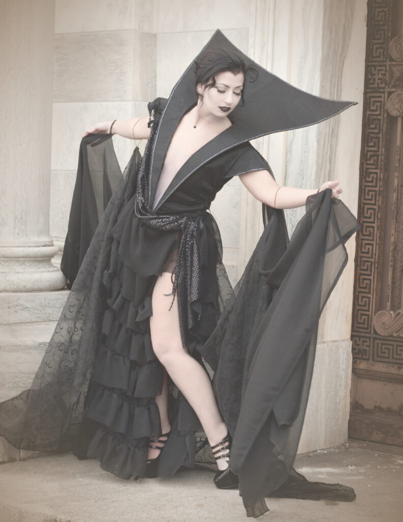 Woman in a long black gown and cape posing on the steps of an ornate stone building.