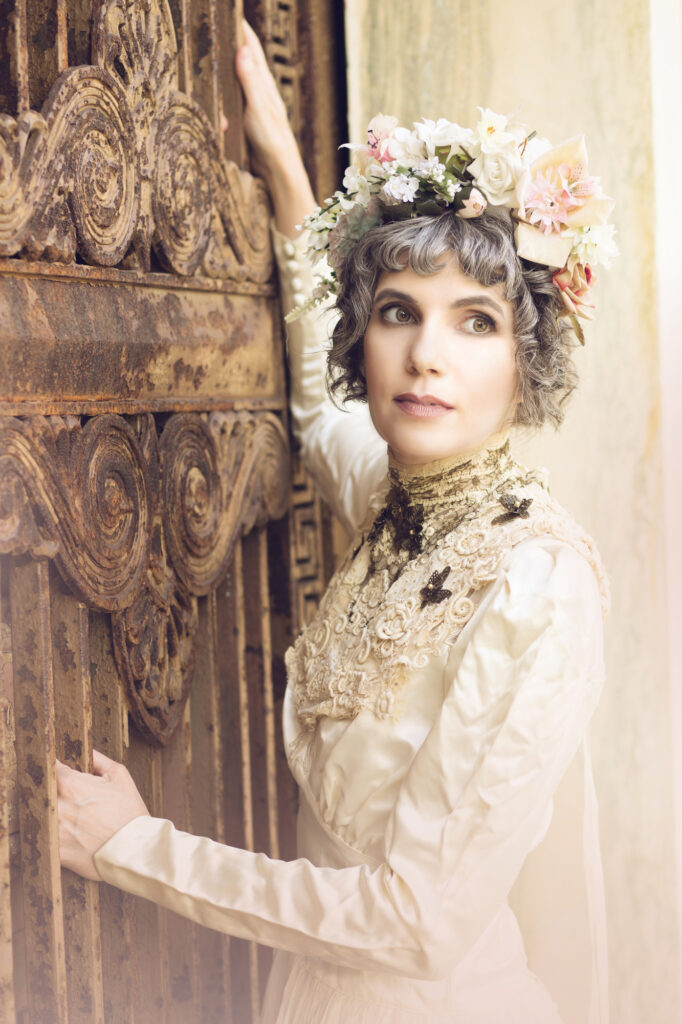 Woman in an ivory Victorian dress and floral headpiece beside an ornate carved doorway.
