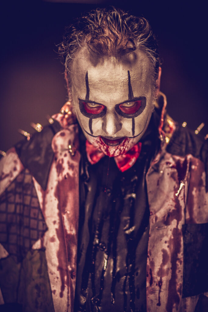Close-up portrait of a blood-covered horror clown with intense red-rimmed eyes.