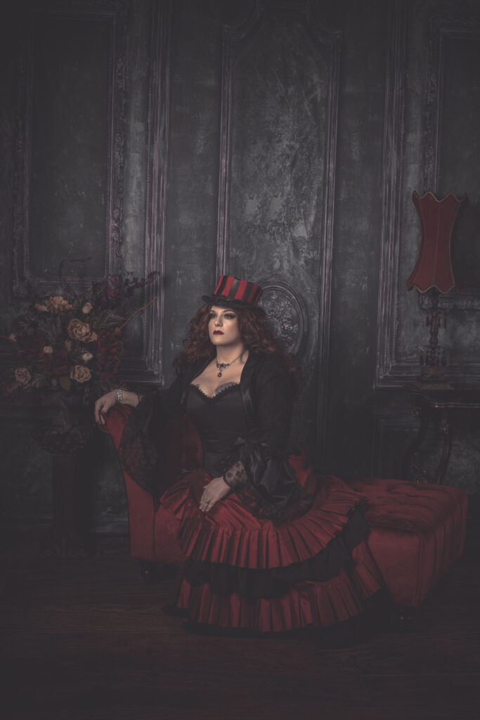 Woman in a red and black steampunk ensemble and striped top hat in a dark parlor.