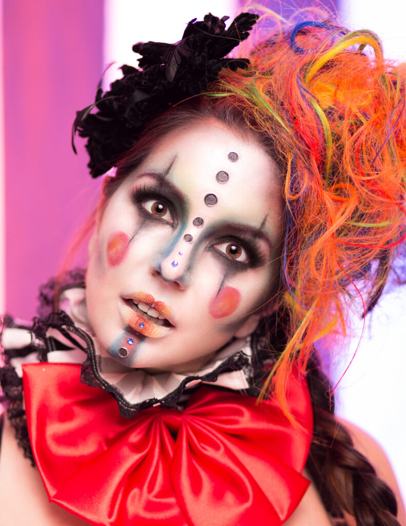 Close-up portrait of a sinister clown with orange hair and colorful graphic makeup.