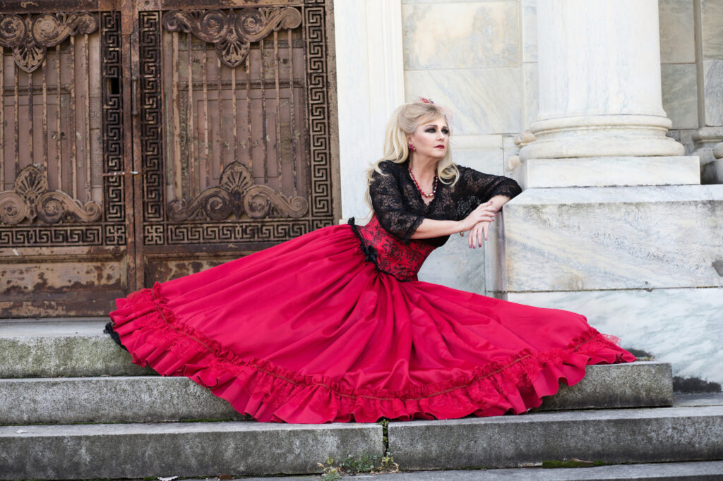 Blonde woman in a black lace top and sweeping red Victorian skirt on stone steps.
