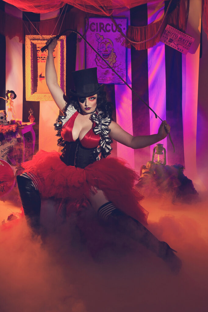 Female magician in a black top hat and red corset inside a colorful theatrical set.