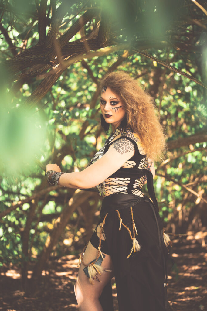 Red-haired fantasy warrior with dramatic eye makeup posing in a sunlit forest.